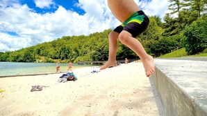 A child jumping off a concrete ledge at a beach with a forested hill in the background. - Hilltop Hideaway- Cozy Cabin w/ Fireplace, Private Deck/Yard, Family Friendly! (Cashiers)