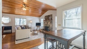 A spacious living room with a wooden ceiling, a stone fireplace, a large window, and a dining area with a wooden table and bar stools. - Hilltop Hideaway- Cozy Cabin w/ Fireplace, Private Deck/Yard, Family Friendly! (Cashiers)