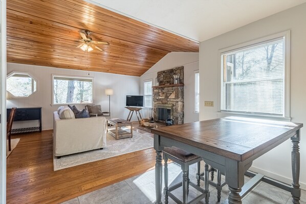 A spacious living room with a wooden ceiling, a stone fireplace, a large window, and a dining area with a wooden table and bar stools.