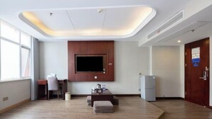 Quarto de hotel moderno com televisão de tela plana na parede, um pequeno espaço de estar com mesa e cadeiras, um refrigerador e uma plataforma de madeira no centro.