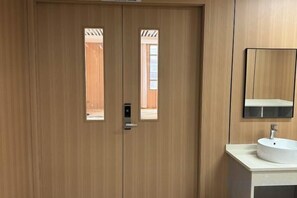 Een houten deur met een sleutelgat en een klein raam, een witte wastafel met een spiegel erboven en uitzicht op het buitenlands landschap door het raam.