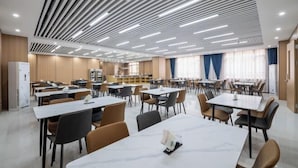 A modern dining area with multiple tables and chairs, a kitchen in the background, and a ceiling with linear lighting fixtures. - Laiyue Hotel (Huidong)