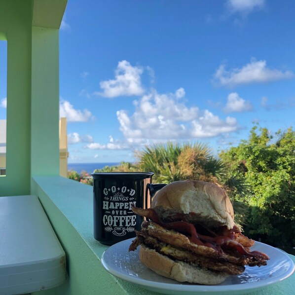 A sandwich on a plate with a coffee mug beside it, set against a backdrop of a clear sky and greenery.