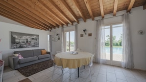 A spacious living area with a round table, a sofa, and a view of a pool through large windows.