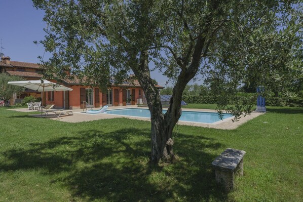 Una villa con piscina, un patio con mobiliario exterior y un gran árbol en primer plano.