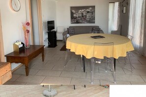 Ein Essbereich mit einem runden Tisch, der gelb bedeckt ist und von transparenten Stühlen umgeben ist. Ein Holztischchen und eine Vase mit Blumen sind zu sehen.