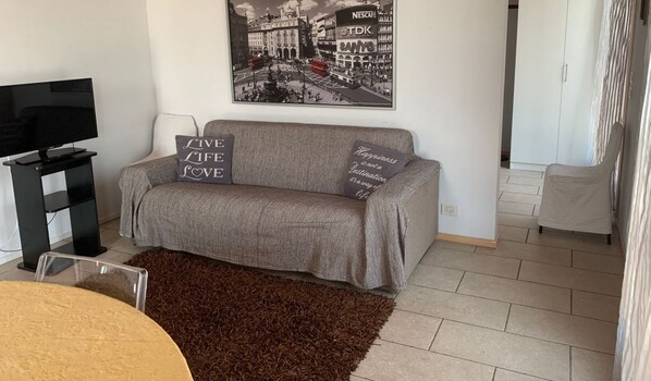 A living room with a grey sofa, a television on a stand, a coffee table, and a brown rug. - La Casina Apartment - with garden and pool (Pozzolengo)