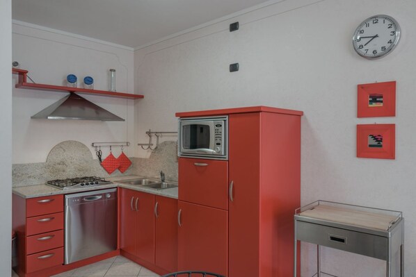 A compact kitchen with red cabinets, a stainless steel dishwasher, a gas stove, a microwave, and a small table with a drawer.