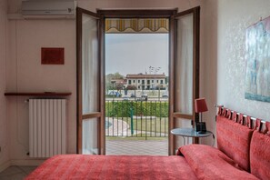 Camera d'albergo con un letto, un termosifone, un tavolino con una lampada e vista sulla strada e sugli edifici circostanti.
