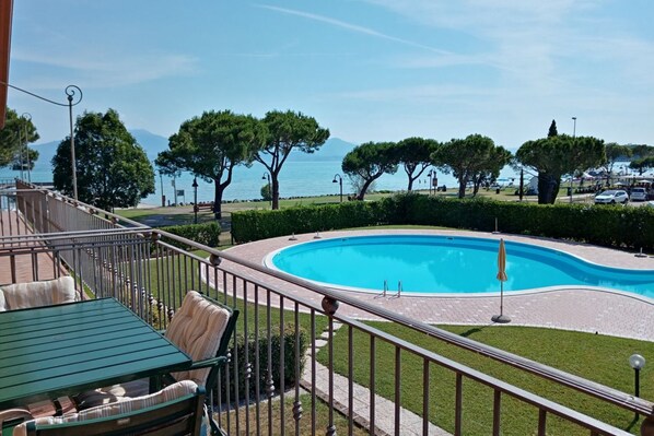 A pool area with a curved design, surrounded by well-maintained hedges and trees, and a view of the sea and mountains in the background.