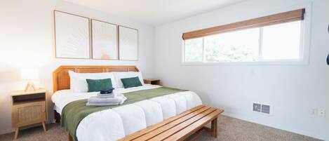 A bedroom with a bed, a wooden bench, a nightstand, a wall-mounted lamp, and a window with blinds.