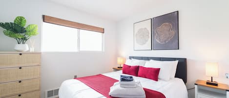 A modern bedroom with a large bed, a bedside table, a dresser, and a window with blinds.