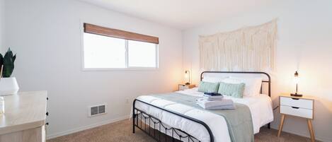 A bedroom with a bed, bedside tables, a window with blinds, a ceiling light, and a wall-mounted macrame piece.