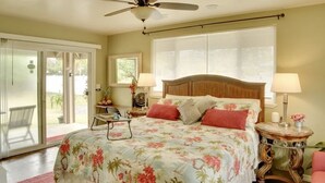 Une chambre à coucher comprenant un lit, deux lampes de chevet, un ventilateur de plafond, une fenêtre avec des stores et une porte-fenêtre coulissante donnant sur un espace extérieur.
