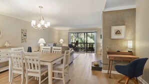 A spacious living area with a dining table, chairs, a sofa, and a desk with a lamp. There is a large window with a view of greenery outside. - 103 Golfmaster- 2 Bedroom Townhouse in Shipyard Ocean Front Community. Golf & Lagoon View Sleeps 6 (Hilton Head Island)