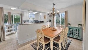 Un espace repas avec une table et des chaises en bois, un lustre, et une vue sur la cuisine et l’espace extérieur.