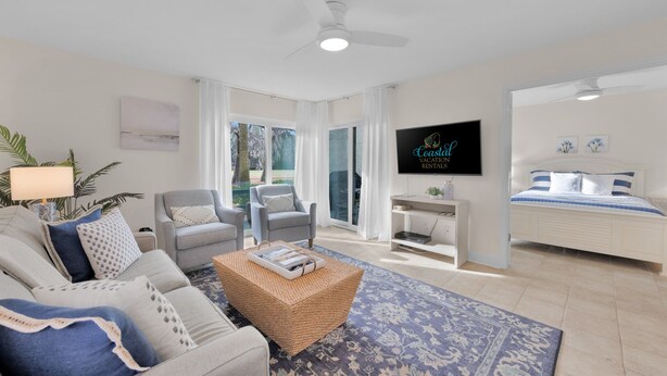 Un salon moderne avec un canapé, des fauteuils, une table basse et un téléviseur. Il y a un balcon avec vue sur les arbres et un ventilateur de plafond.