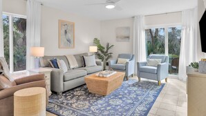 Un salon moderne avec un canapé gris, un fauteuil bleu, une table basse en osier et un tapis à motifs. On y trouve un ventilateur de plafond, un tableau au mur et une vue sur la verdure à travers les fenêtres.