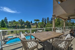 A patio with a wooden table and wicker chairs, overlooking a swimming pool and a lake with trees and houses in the background. - Luxe Getaway w/ Riverfront Views in Grants Pass! (Grants Pass)