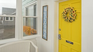 A bright yellow door with a sunflower wreath, a 'WELCOME' sign, and a bench in a hallway with wooden flooring. - SB2-Downtown Munising, Deli and Brewery Across the Street (Munising)