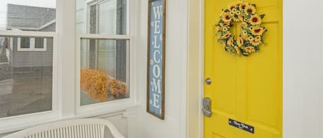 Uma porta amarela vibrante adornada com um arranjo de girassóis, um letreiro com a palavra 'WELCOME' e um banco em um corredor com piso de madeira.
