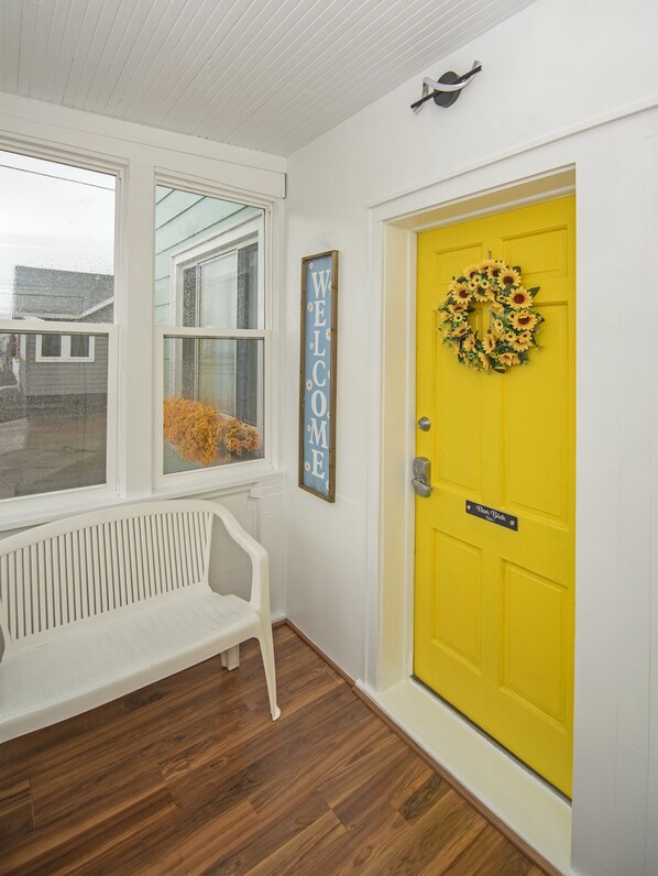 Una puerta amarilla brillante con un arreglo de girasoles, un letrero que dice 'WELCOME' y un banco en un pasillo con piso de madera.