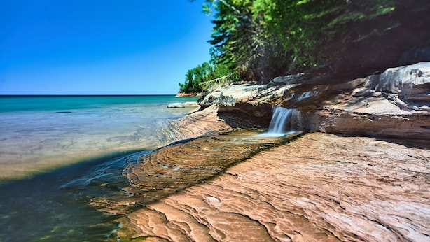 A clear view of a rocky shoreline with a waterfall, calm water, and lush greenery. - SB2-Downtown Munising, Deli and Brewery Across the Street (Munising)