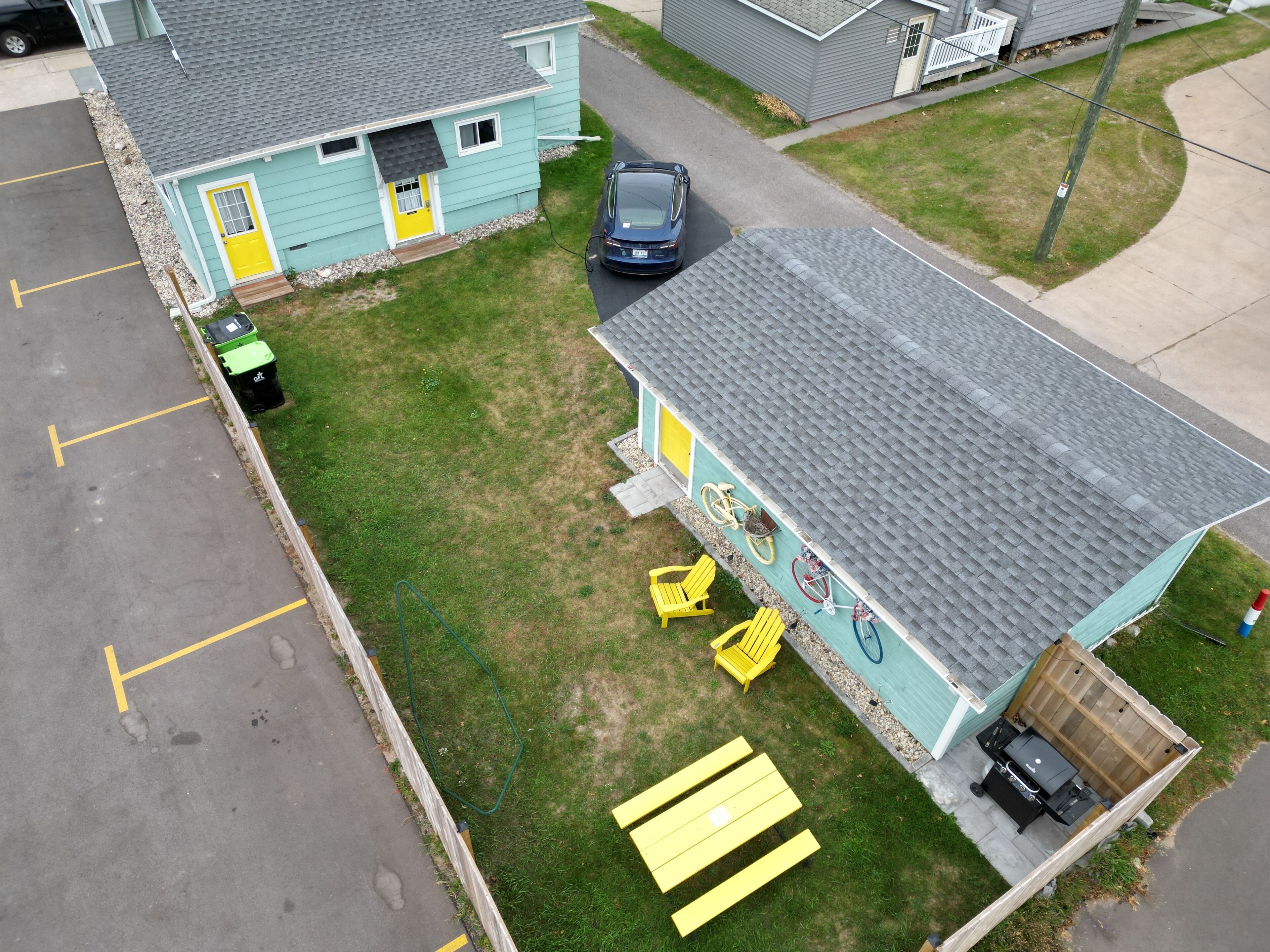 Un petit bâtiment d’un seul étage, à l’extérieur bleu et à la porte jaune. Une voiture est garée devant, ainsi qu’une tondeuse à gazon verte et un vélo appuyé contre le bâtiment.