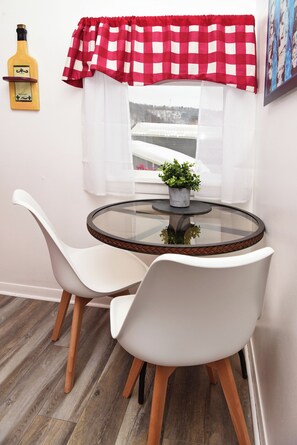 A round glass table with two white chairs, a small potted plant, a bottle of liquor on a wall-mounted shelf, and a window with a red and white checkered curtain.