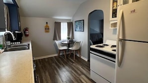 A compact kitchen with a white refrigerator, stove, and sink. There's a dining area with a table and chairs, and a window with curtains. - TH3-Updated Apartment Downtown Munising, Walk to Attractions & Lake Superior (Munising)