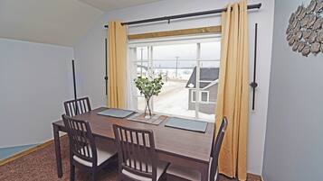 A dining area with a wooden table, four chairs, a vase with a plant, and a view of a snowy landscape through a window with yellow curtains.