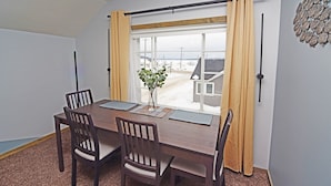 A dining area with a wooden table, four chairs, a vase with a plant, and a view of a snowy landscape through a window with yellow curtains. - TH3-Updated Apartment Downtown Munising, Walk to Attractions & Lake Superior (Munising)