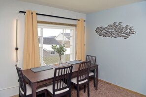 Un rincón comedor con una mesa y sillas, una ventana con cortinas, un florero con planta y una obra de arte en la pared.