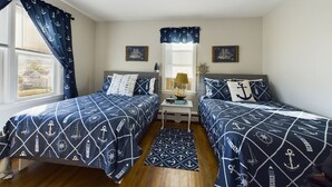 Une chambre avec deux lits, de la literie sur le thème de la marine, une petite table avec une lampe et une fenêtre avec des rideaux.
