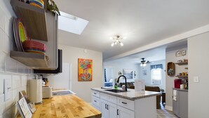 Une cuisine moderne avec des armoires blanches, un comptoir en bois et un îlot central. Un tableau coloré orne le mur, ainsi qu’un ventilateur de plafond et un panneau indiquant un bar à café.
