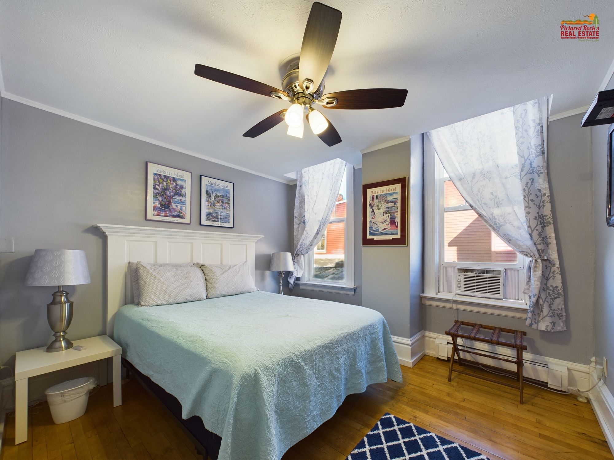 A bedroom with a bed, a ceiling fan, a window with curtains, and framed pictures on the wall.