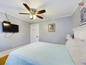 Une chambre à coucher avec un lit, un ventilateur de plafond, un téléviseur fixé au mur, une porte et un tableau encadré accroché au mur.