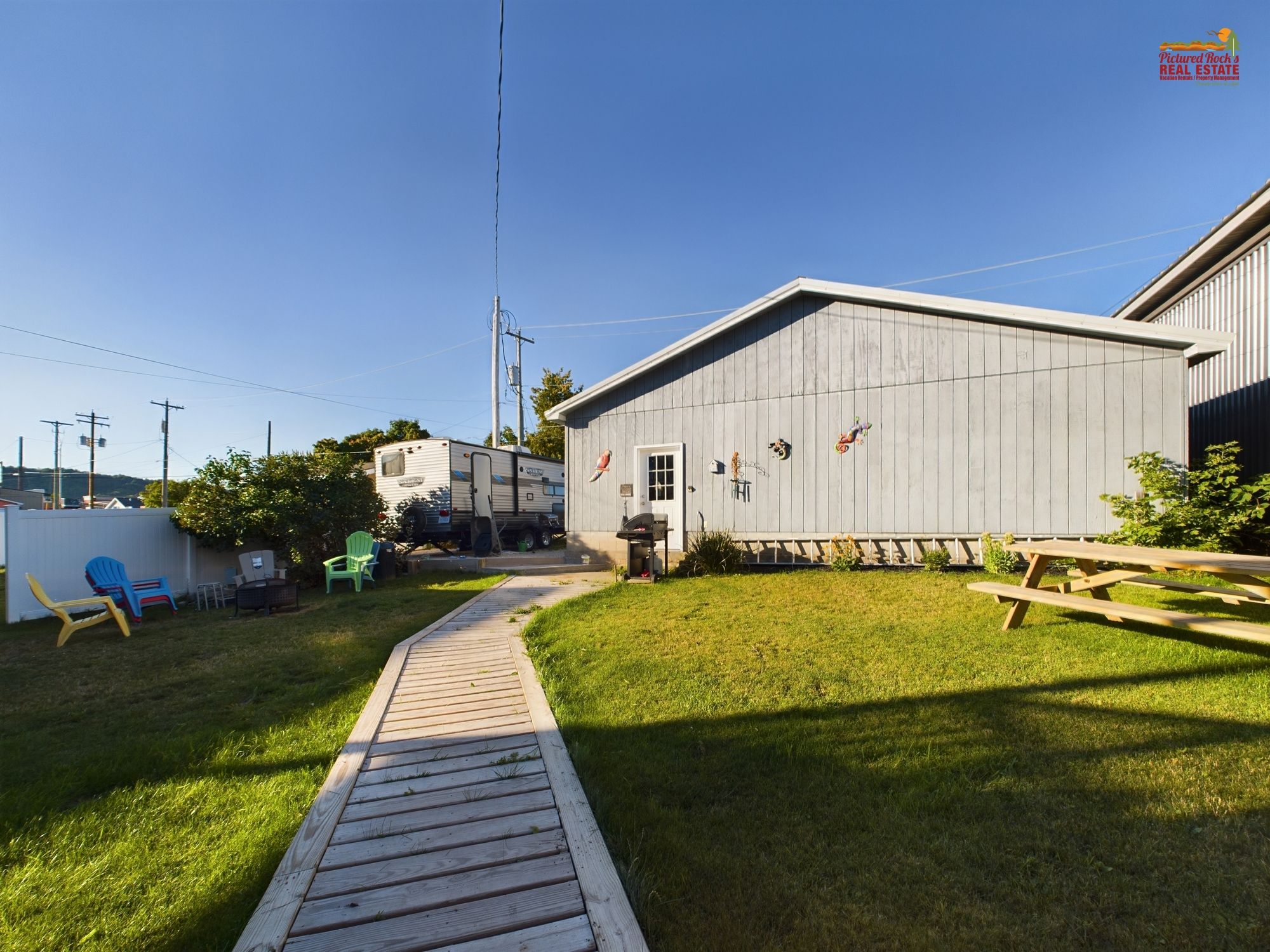 A backyard with a wooden deck leading to a building, a picnic table, and a camper parked nearby.