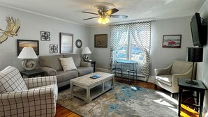 A living room with a wooden floor, a ceiling fan, a sofa, a coffee table, and a dining area with a table and chairs. - SSVR2-Spacious, Downtown Munising Apartment (Munising)