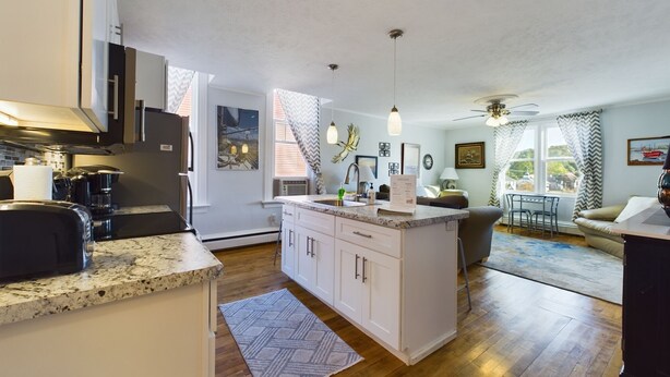 A modern kitchen with granite countertops, white cabinetry, and a central island. There is a dining area with a table and chairs, a sofa, and a view of the outdoors through large windows. - SSVR2-Spacious, Downtown Munising Apartment (Munising)