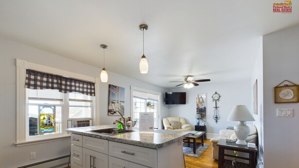 A modern kitchen with a large island, pendant lights, and a ceiling fan. There is a dining area with a table and chairs, and a living room with a sofa and a lamp.