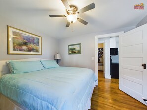 A bedroom with a bed, a ceiling fan, a painting, and a door leading to a kitchen.