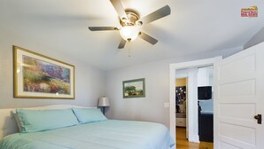A bedroom with a bed, a ceiling fan, a painting, and a door leading to a kitchen.