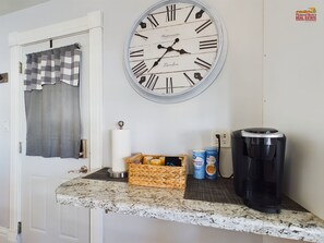 A kitchen with a large wall clock, a coffee maker, and a basket of snacks.