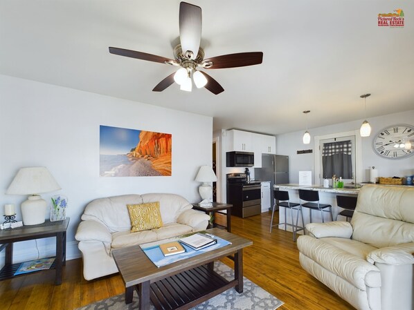 A modern living room with a sofa, a coffee table, and a ceiling fan. There is a kitchen area with a refrigerator and a dining table.