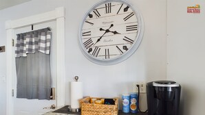 A kitchen with a large wall clock, a coffee maker, and a basket of snacks.