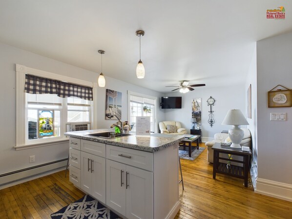 A modern kitchen with a large island, pendant lights, and a ceiling fan. There is a dining area with a table and chairs, and a living room with a sofa and a lamp.