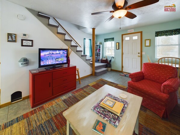 A living room with a staircase, a television, a red sofa, a coffee table, and a bookshelf. - Rock House-Downtown Munising with Private Patio, Garage, Fire-pit & BBQ Grill (Munising)
