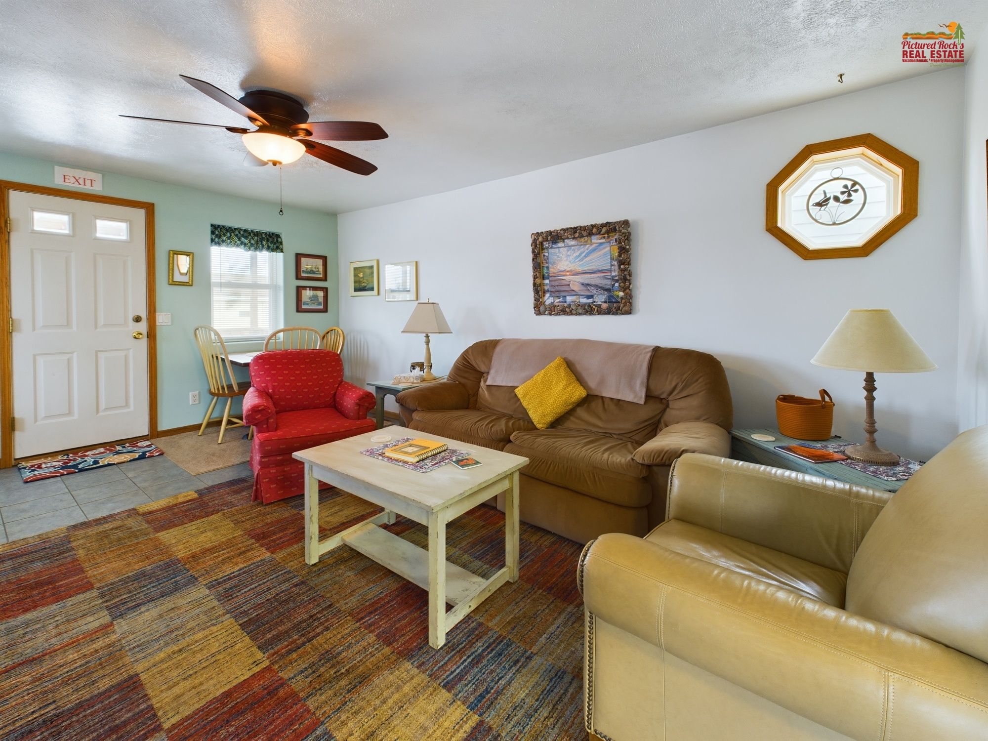 A living room with a ceiling fan, a brown sofa, a red armchair, a beige armchair, a coffee table, and a patterned rug.