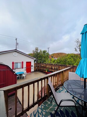 Eine überdachte Terrasse mit Tisch und Stühlen, ein rotes Gebäude im Hintergrund und ein Holzzaun.